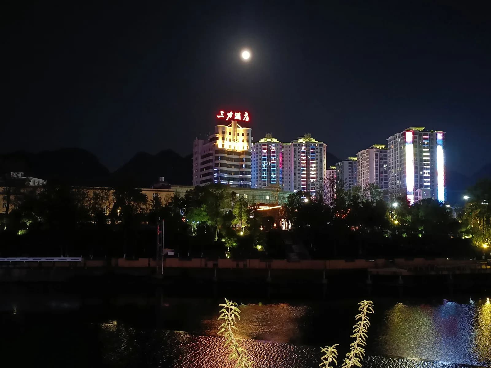 Night skyline of Libo county town in Guizhou with lit high-rise buildings above the riverfront under a full moon