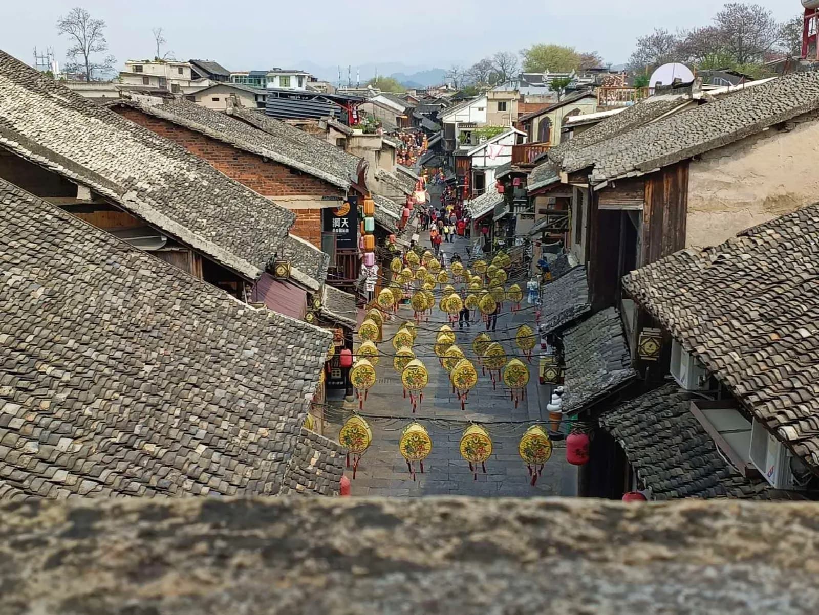 View from the Qingyan city wall over grey-tiled rooftops and a lantern-lined street in March 2024