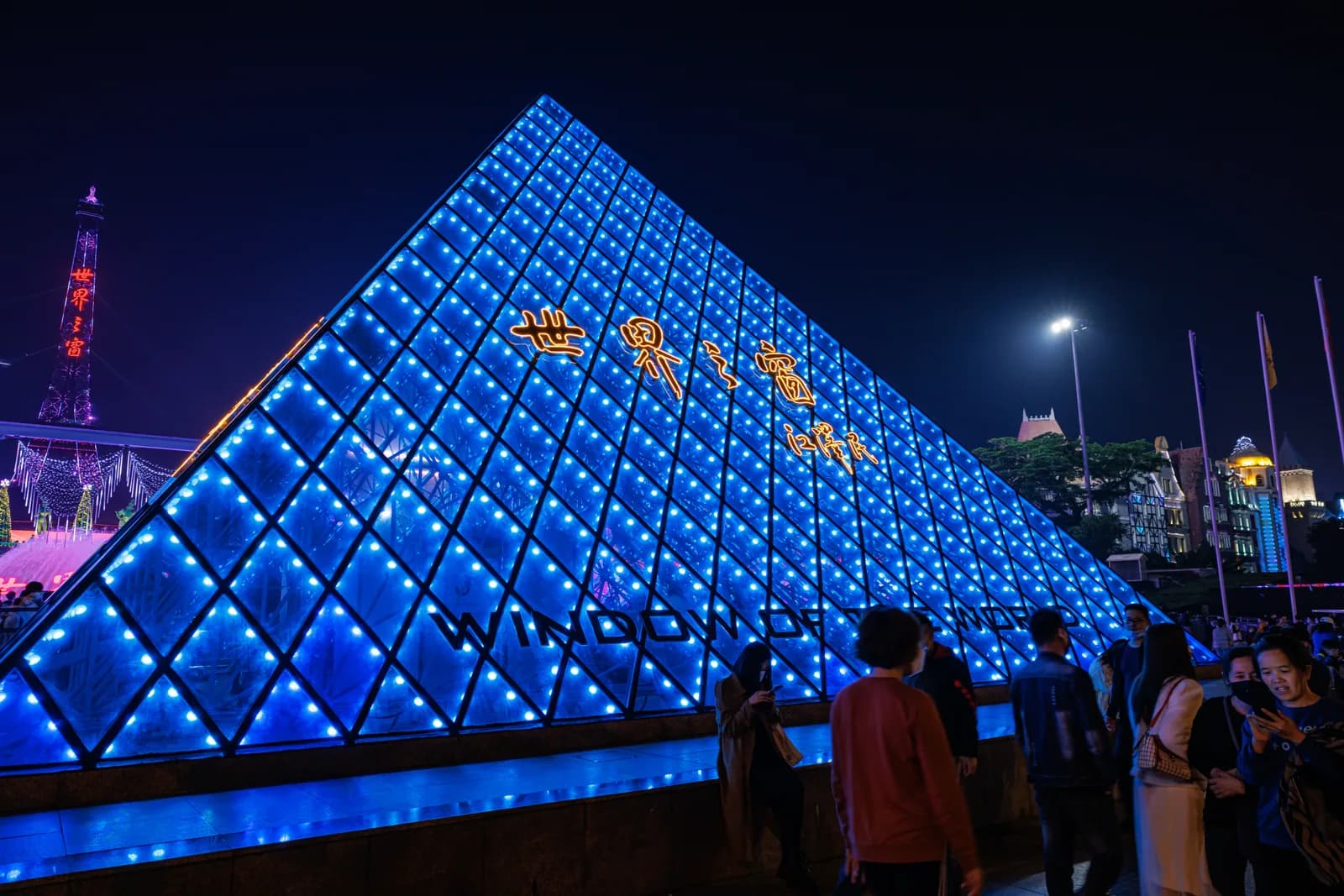The illuminated blue glass pyramid at the entrance of Window of the World in Shenzhen at night with the lit Eiffel Tower replica behind
