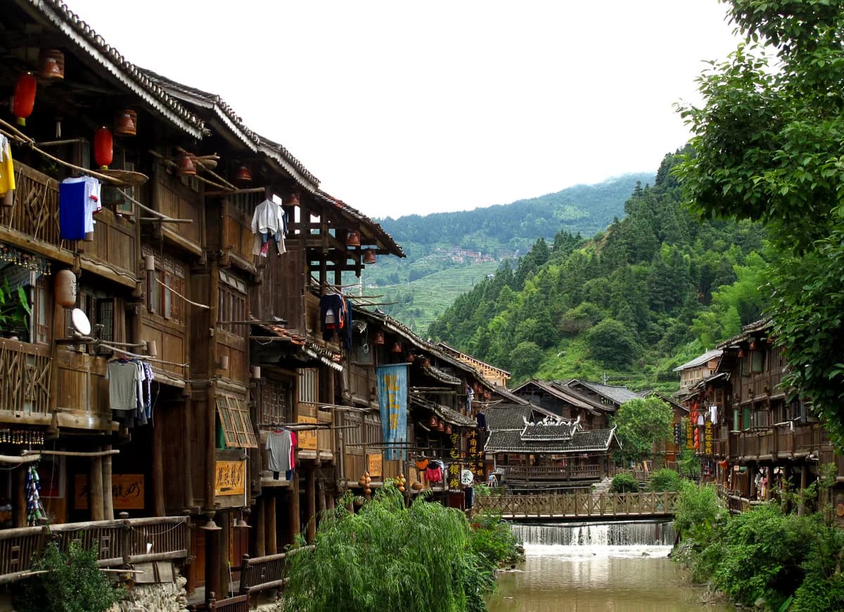 Traditional wooden Dong houses lining the stream through Zhaoxing village, with a wind-and-rain bridge in the distance