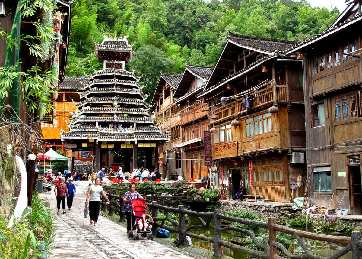 A multi-tiered wooden drum tower rising over a busy stone-paved lane in Zhaoxing Dong village, with shops and visitors below