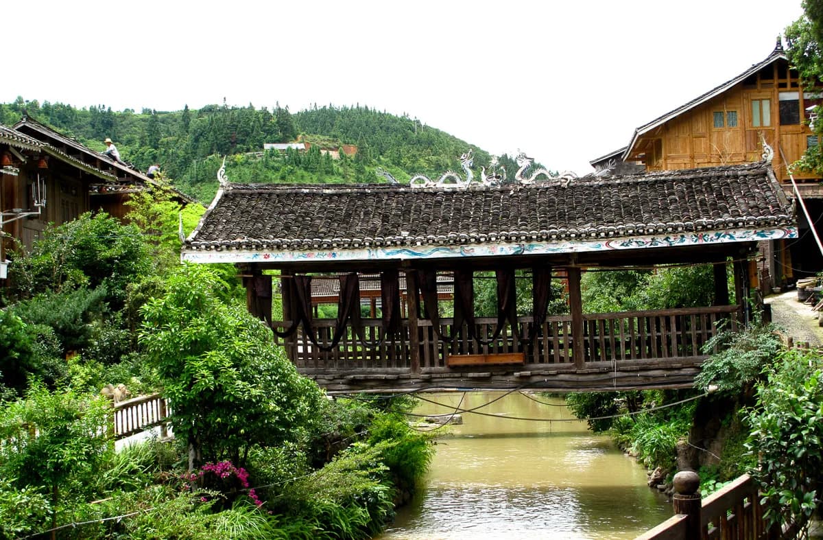 A covered wooden wind-and-rain bridge with a tiled roof crossing the stream in Zhaoxing Dong village, Guizhou